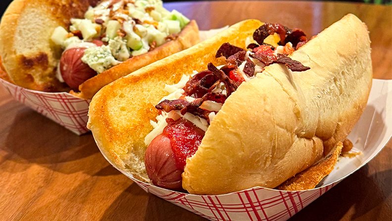 Two loaded hot dogs in takeout containers on wooden table.
