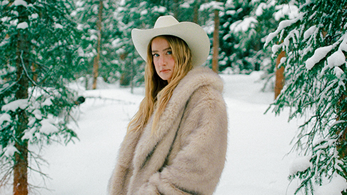 Singer Ingrid Andress in a fur coat and cowboy hat standing in a snowy forest.